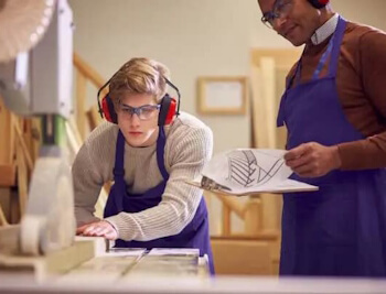 Schreinerlernender in der Lernstufe Berufsfachschule schneidet in der Werkstatt Holz an der Kreissäge zu und wird vom Lehrmeister angeleitet.