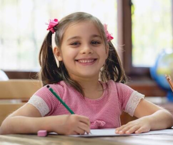 Kind in der Lernstufe Primarschule/2. Zyklus schaut vom Schreiben auf und lacht in die Kamera – Lernen innerhalb der Altersstufen.